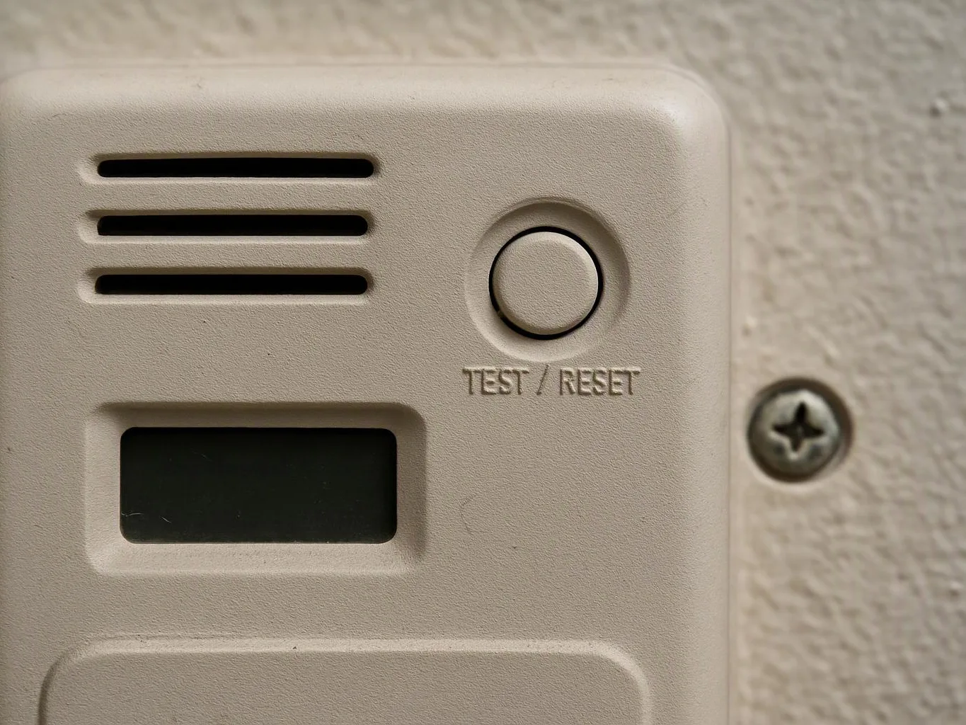A beige wall alarm device shows a TEST RESET button, vents, and a dark display window mounted on textured paint