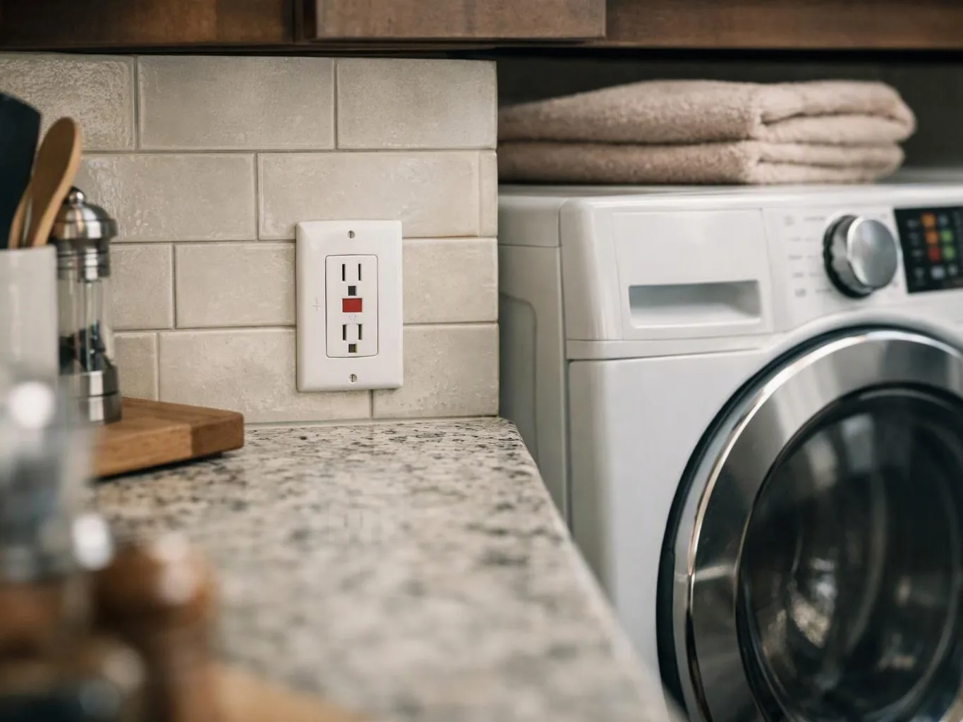 A laundry room counter sits beside a front-loading washer, with folded towels and an outlet on tiled backsplash